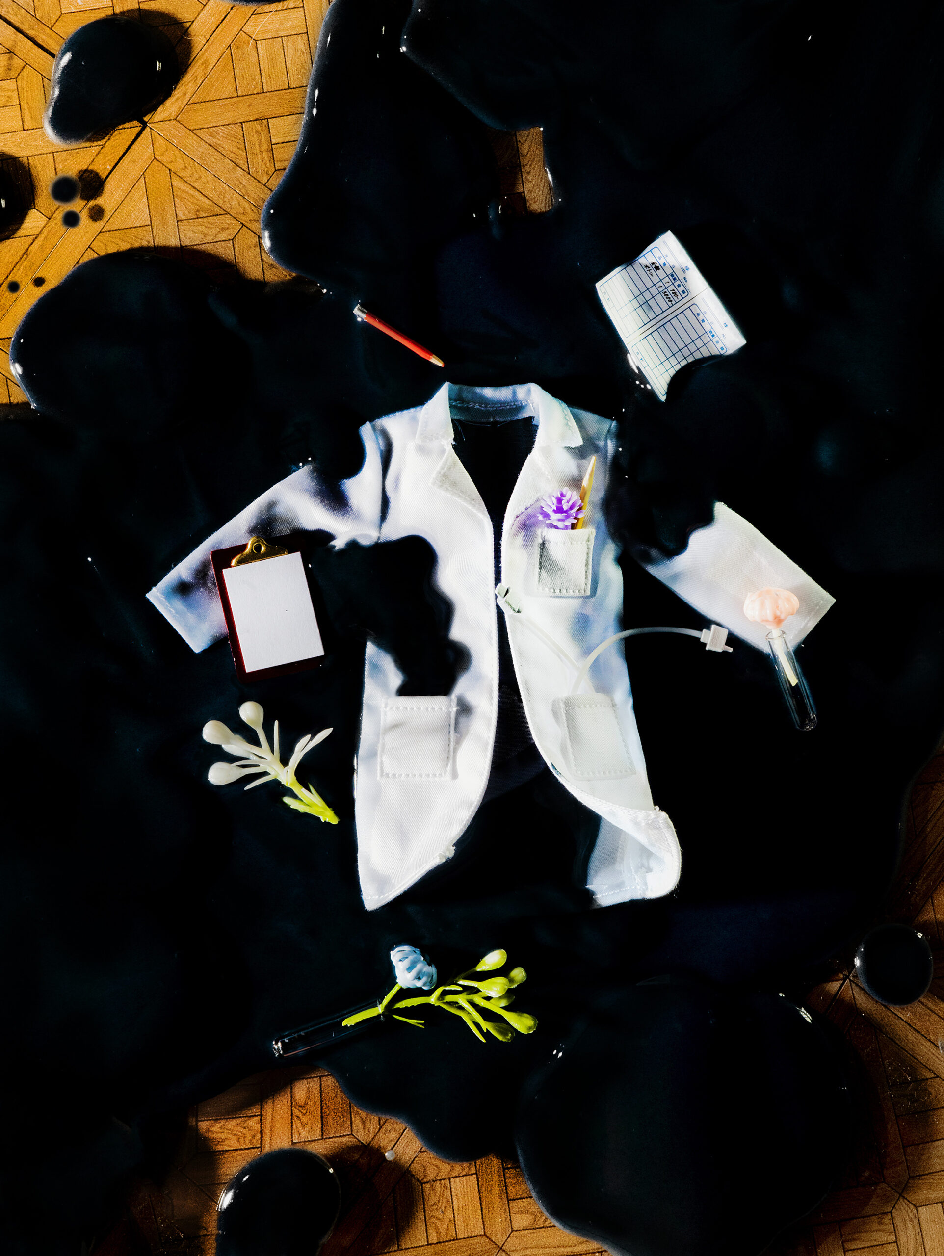 A lab coat surrounded by flowers and office supplies sits in a pool of splattered black oil.
