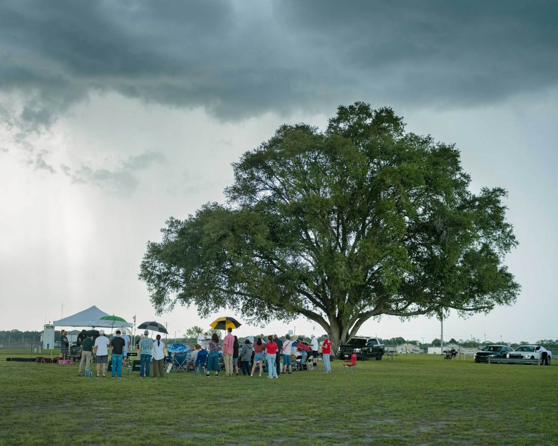 A wide shot captures a large crowd of people gathered on a vast green lawn under a massive, spreading oak tree. Several individuals are holding open umbrellas or standing near lawn chairs and small pop-up canopies, while a law enforcement vehicle is parked to the right of the tree.