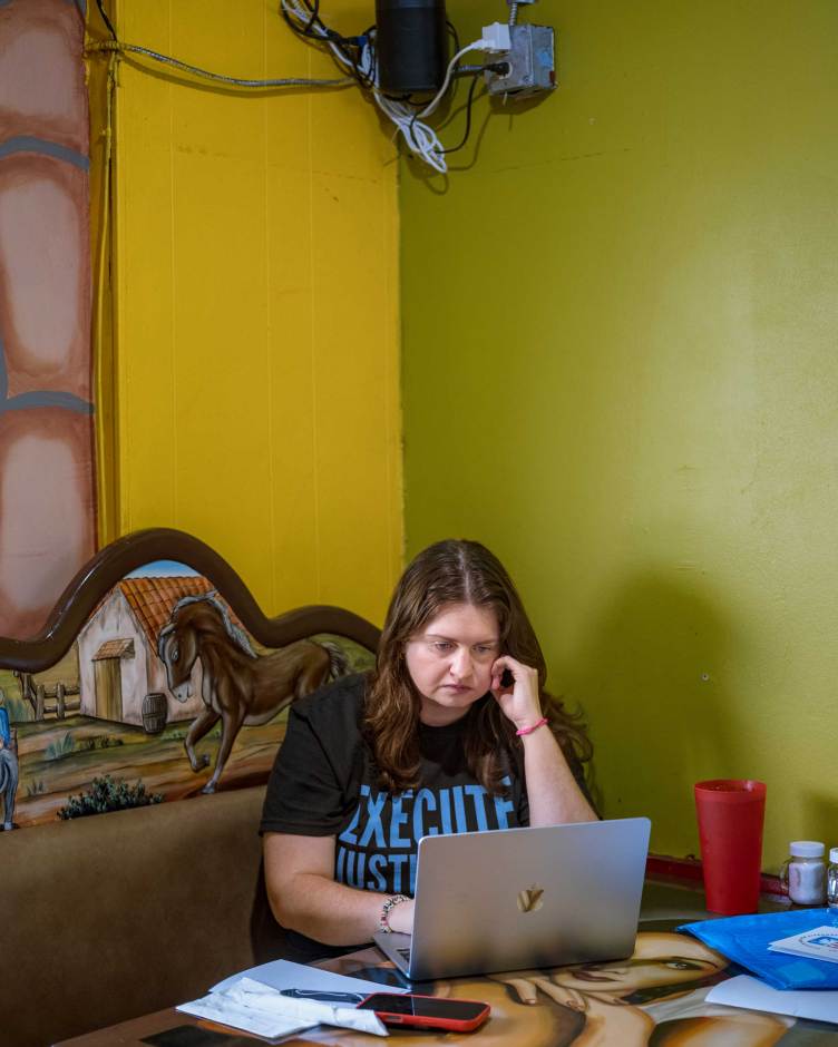 A woman with brown hair sits at a table in the corner of a room with yellow and green walls, looking intently at an open laptop. She is wearing a black T-shirt with blue text that reads “Execute Justice” and resting her chin on her hand.