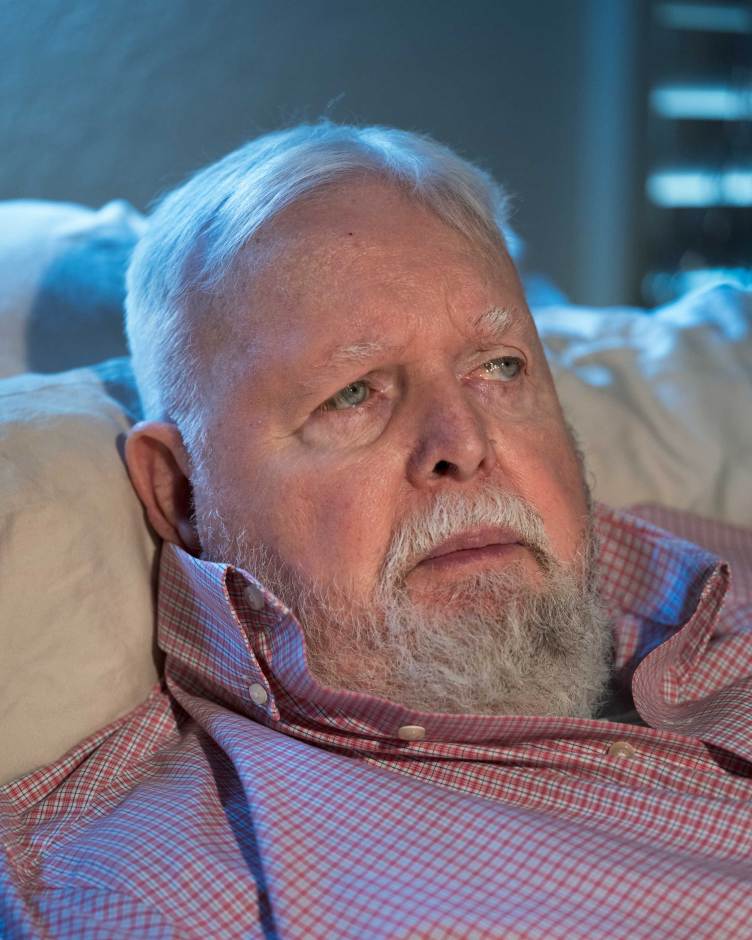 A close-up portrait shows an older man with white hair, a white beard and blue eyes resting against a light-colored pillow.