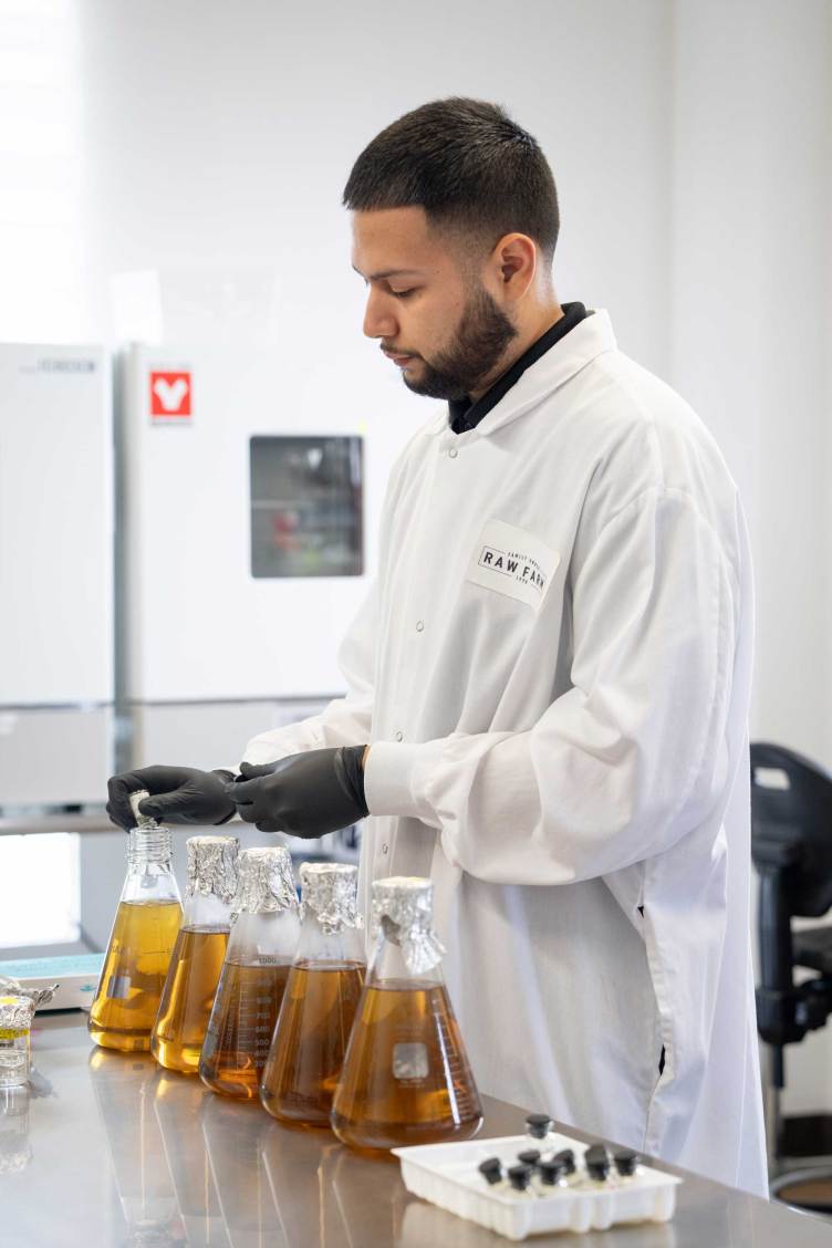 A man in a white lab coat and black gloves works in a laboratory setting. He is handling glass flasks containing an amber liquid lined up on a stainless steel countertop. In the background, lab equipment and a refrigeration unit are visible.