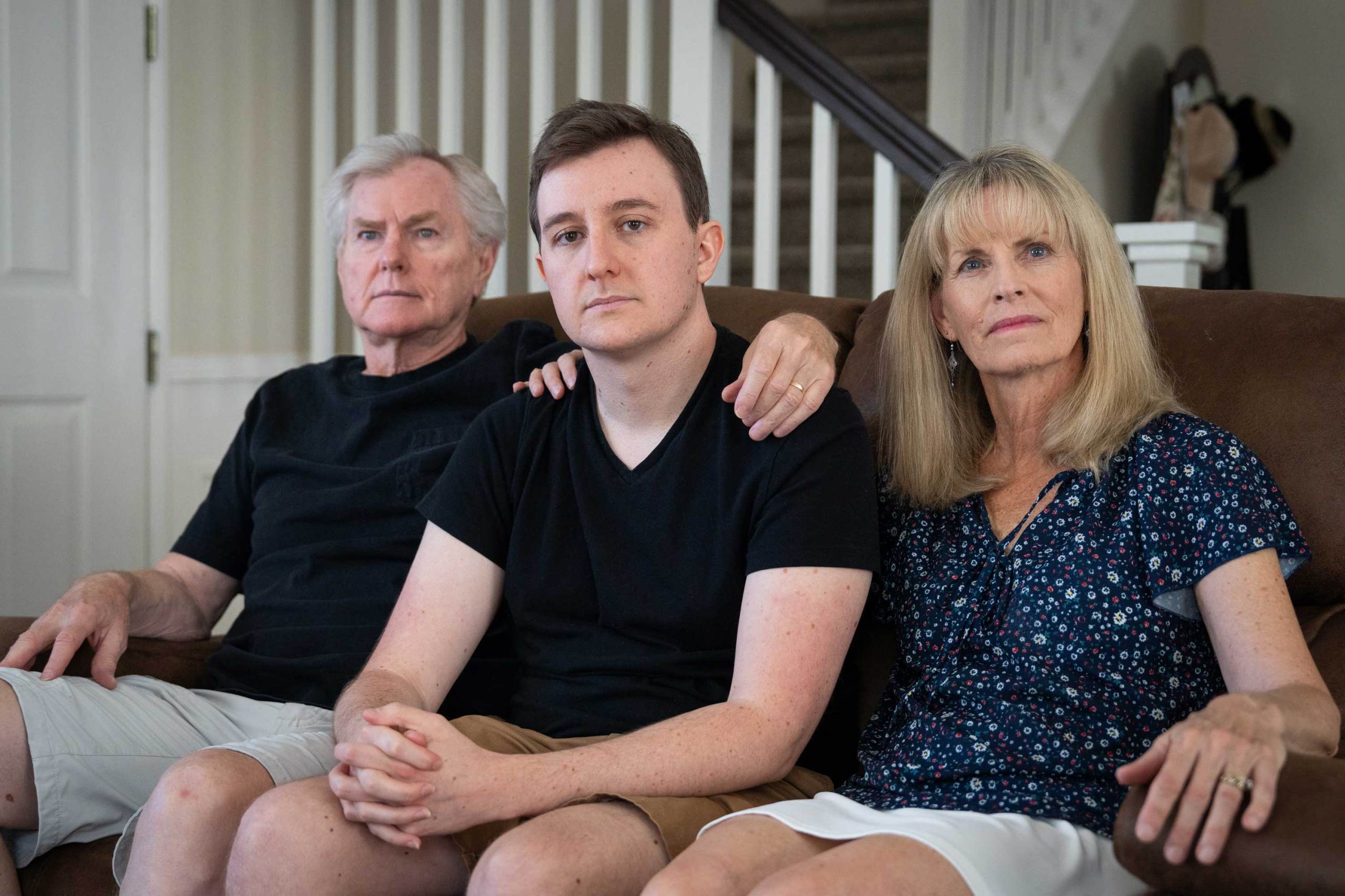 Three people — an older man, a younger man and an older woman — sit together on a brown leather couch in a living room, all wearing serious expressions. The older people rest their hands on the younger man’s shoulders.