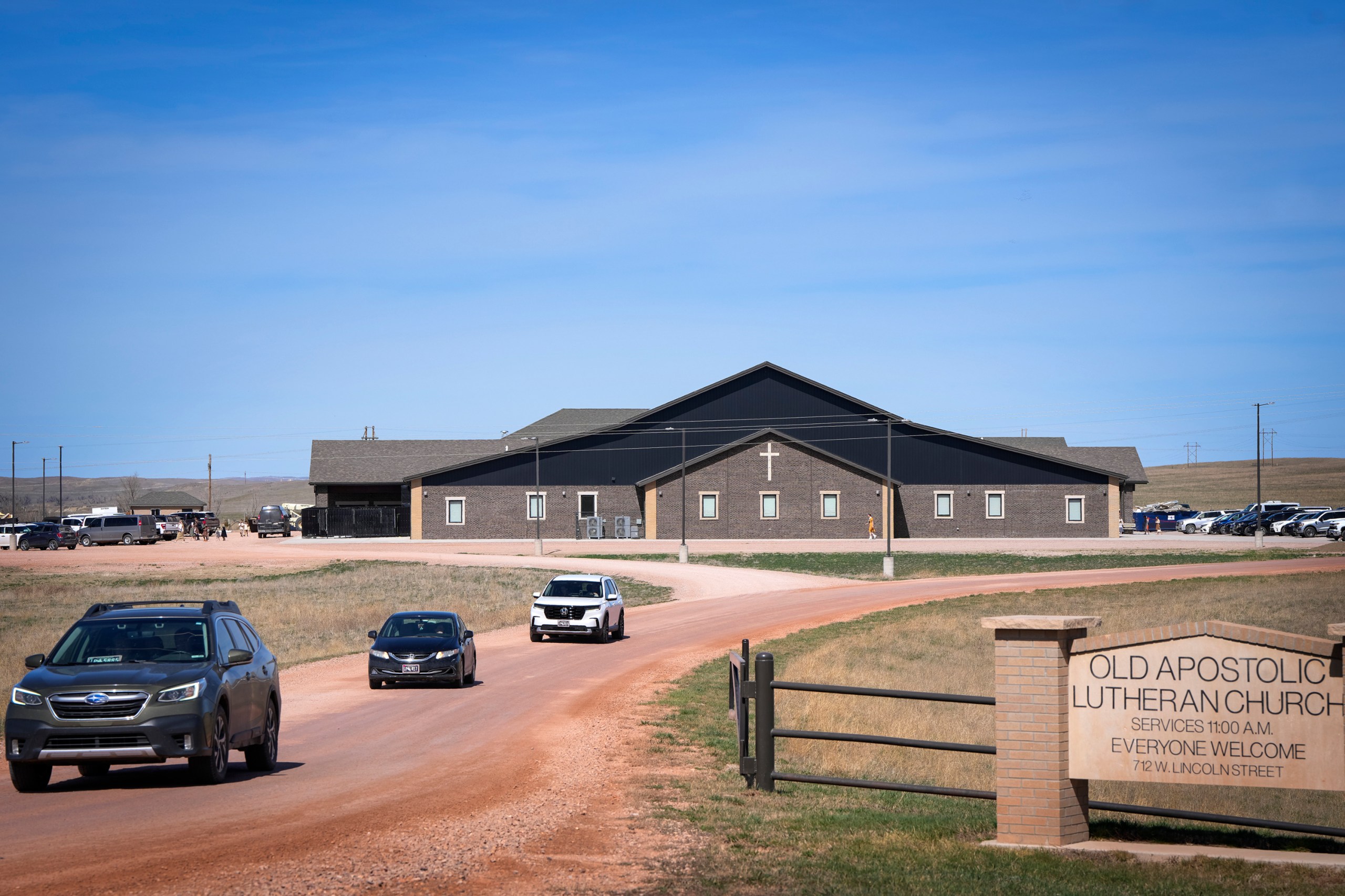 A modern, dark-brick building in a vast, rural landscape under a clear blue sky. A dirt road leads to the church, with a few cars driving on it, and a sign in the foreground says "Old Apostolic Lutheran Church” and “Everyone Welcome."
