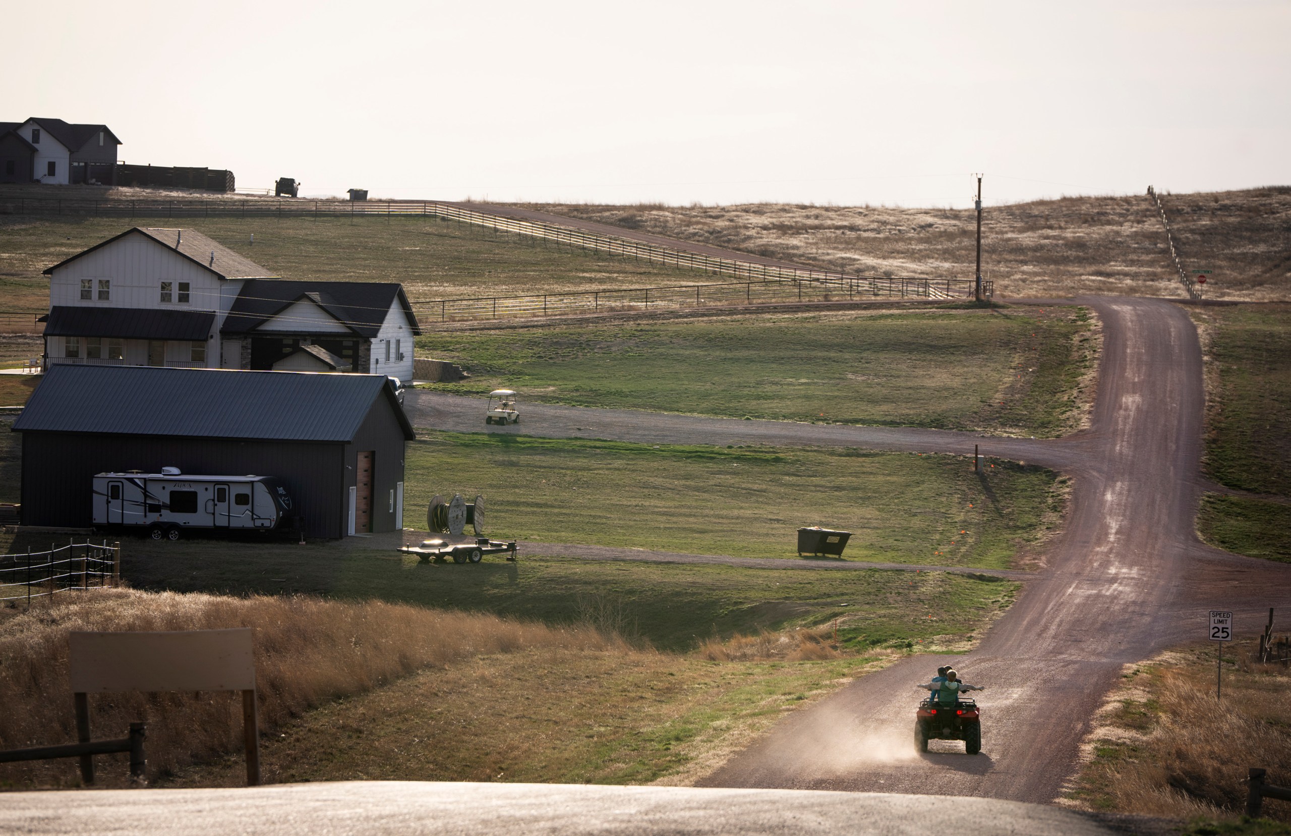A rural area with a few houses, barns, an RV and a dirt road where two people are riding away on an all-terrain vehicle.