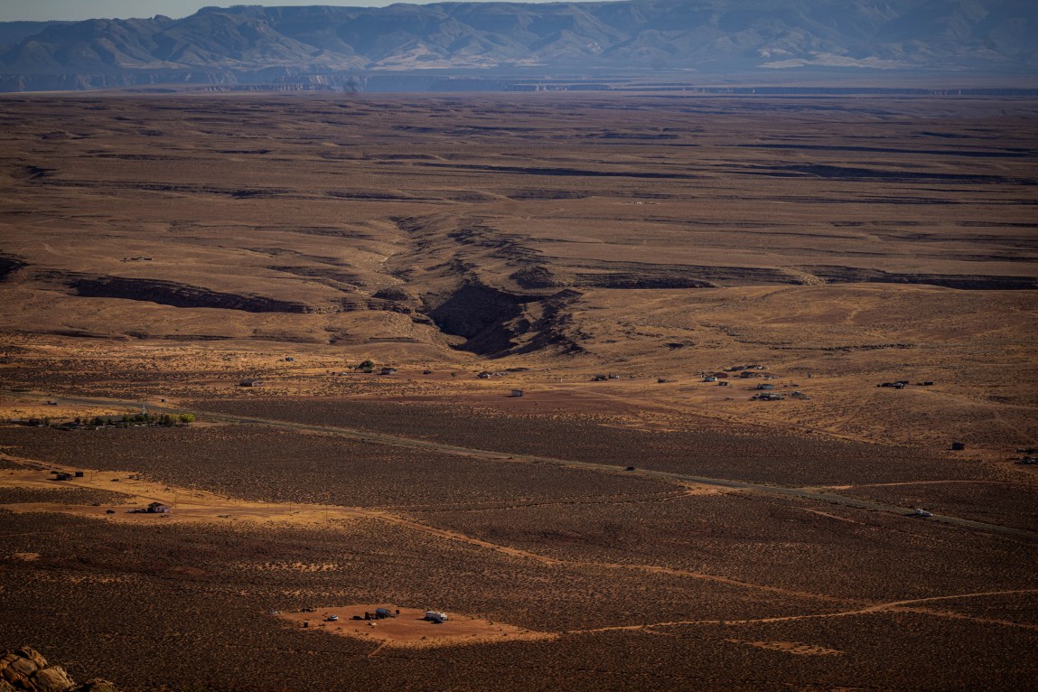 A wide, high-angle view of a vast, arid desert landscape under a hazy sky, with scattered small structures and dirt roads stretching toward a distant mountain range.