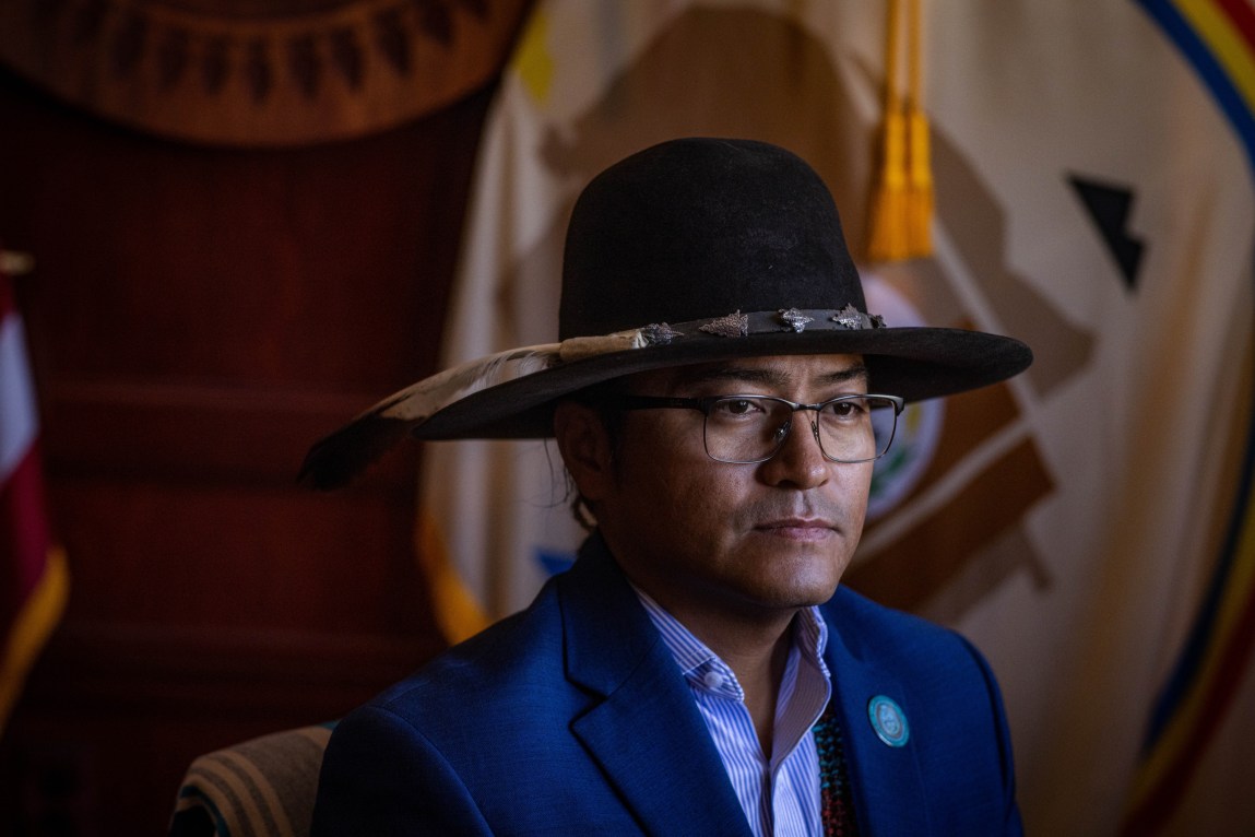 A portrait of a man wearing a blue suit jacket, a striped collared shirt and glasses. He wears a wide-brimmed black hat adorned with silver accents and a feather, and is looking slightly off-camera against a background featuring a tribal flag.