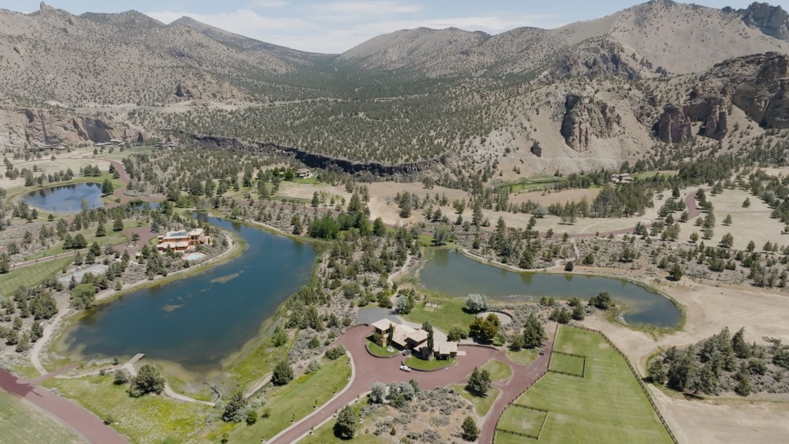 A wide, sweeping landscape photograph showing mansions, large, lush green lawns and pools of water, with mountains in the background.