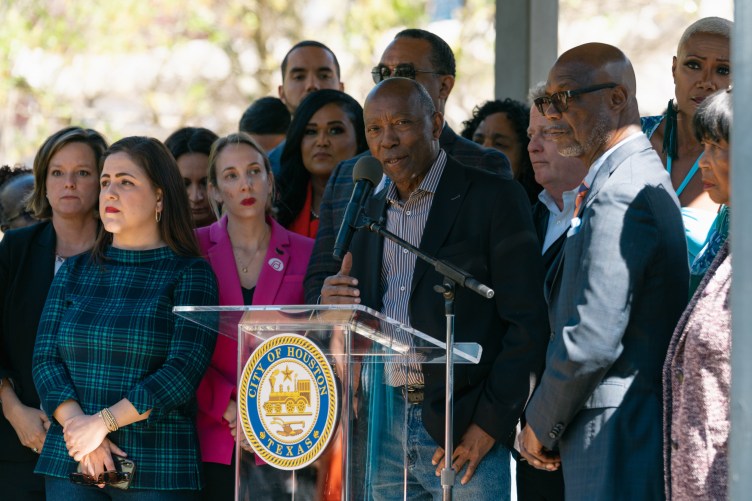 A man speaks at a lectern bearing the city of Houston seal, surrounded by a group of people during an outdoor press conference.
