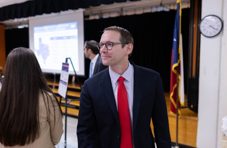 A man wearing a navy suit, glasses and a bright red tie. He is smiling slightly while walking through a meeting at a school, with a projection screen displaying a map of Texas and a Texas state flag visible in the background.
