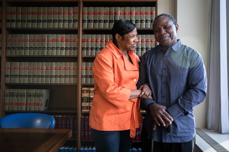 A man with a cane is smiling with his eyes closed while a woman grasps his arm and smiles. Behind them are shelves of books.