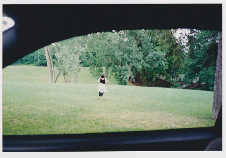 Seen through a car window, a woman walks across a vast lawn with trees behind her.