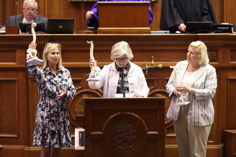 Three women stand at a legislative podium holding up anatomical models of human spines.