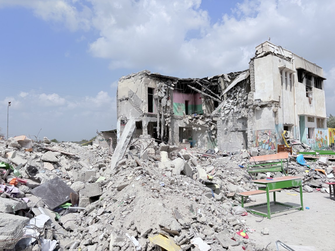 A partially destroyed building, with piles of rubble and school desks in front of it.