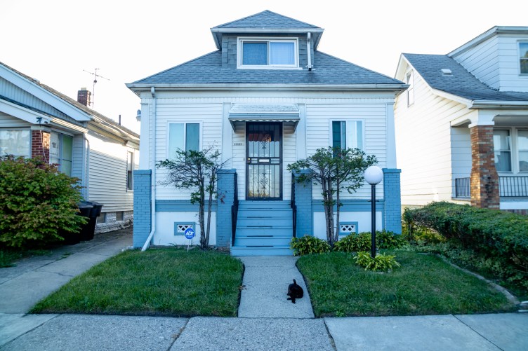 A row of houses, seen from the sidewalk, with a black cat sitting in front of the center house.
