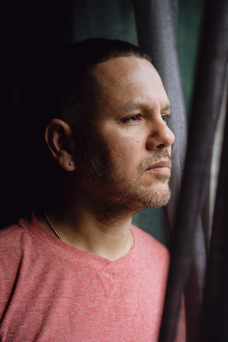 A close-up profile portrait of a man with short hair and light stubble looking thoughtfully out a window beside a dark curtain.