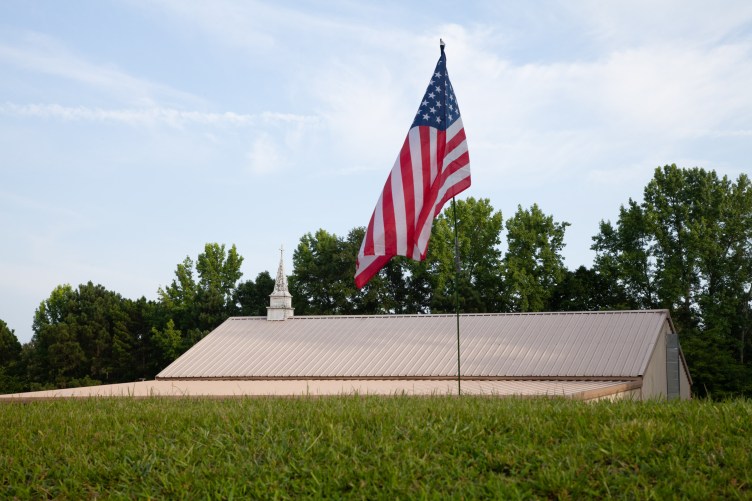 A U.S. flag planted in a green lawn waves in the foreground in front of a white building with a small steeple.