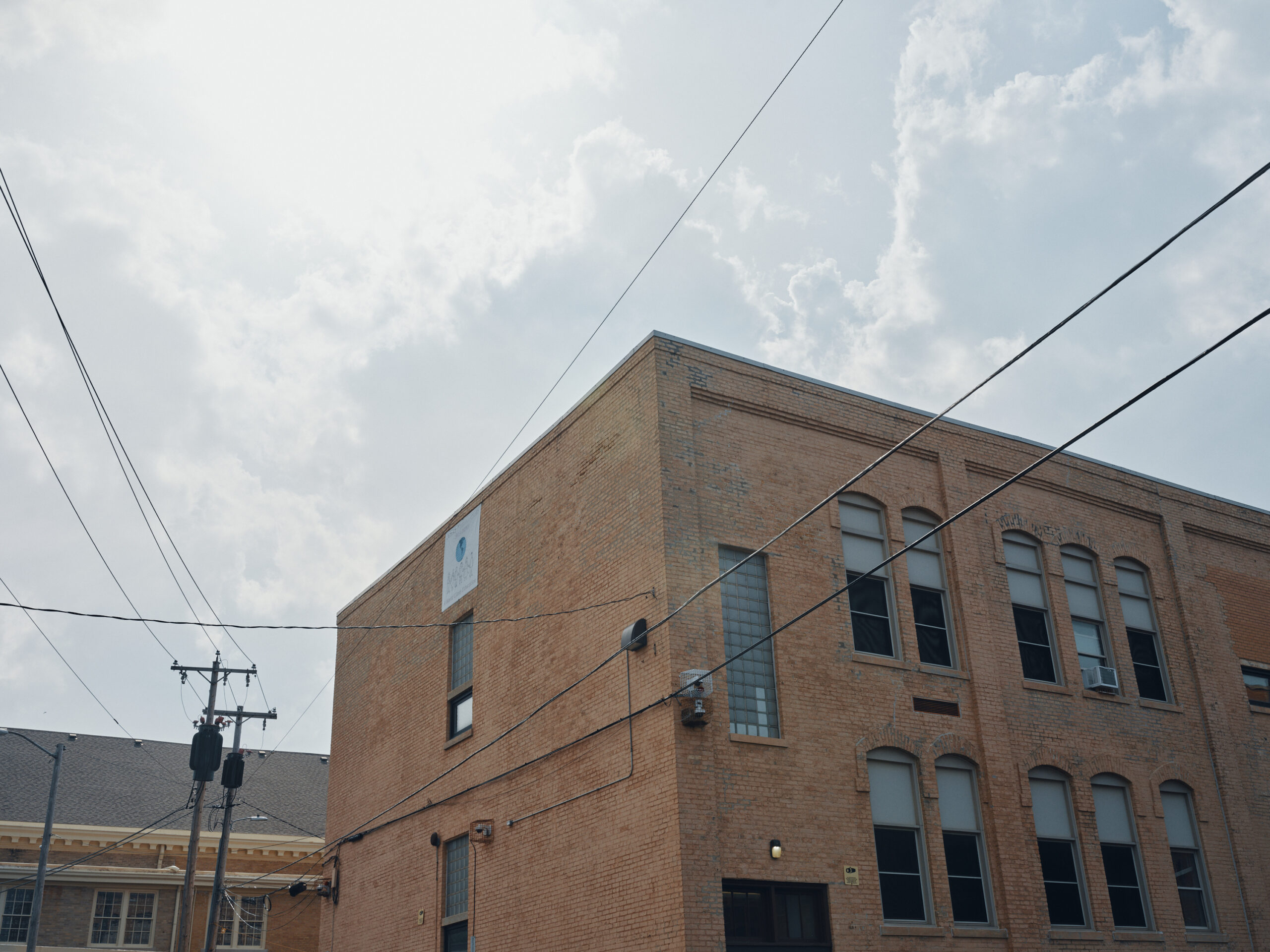 Power lines cross near a two-story, brown brick building with tall windows. The sky has large gray clouds.