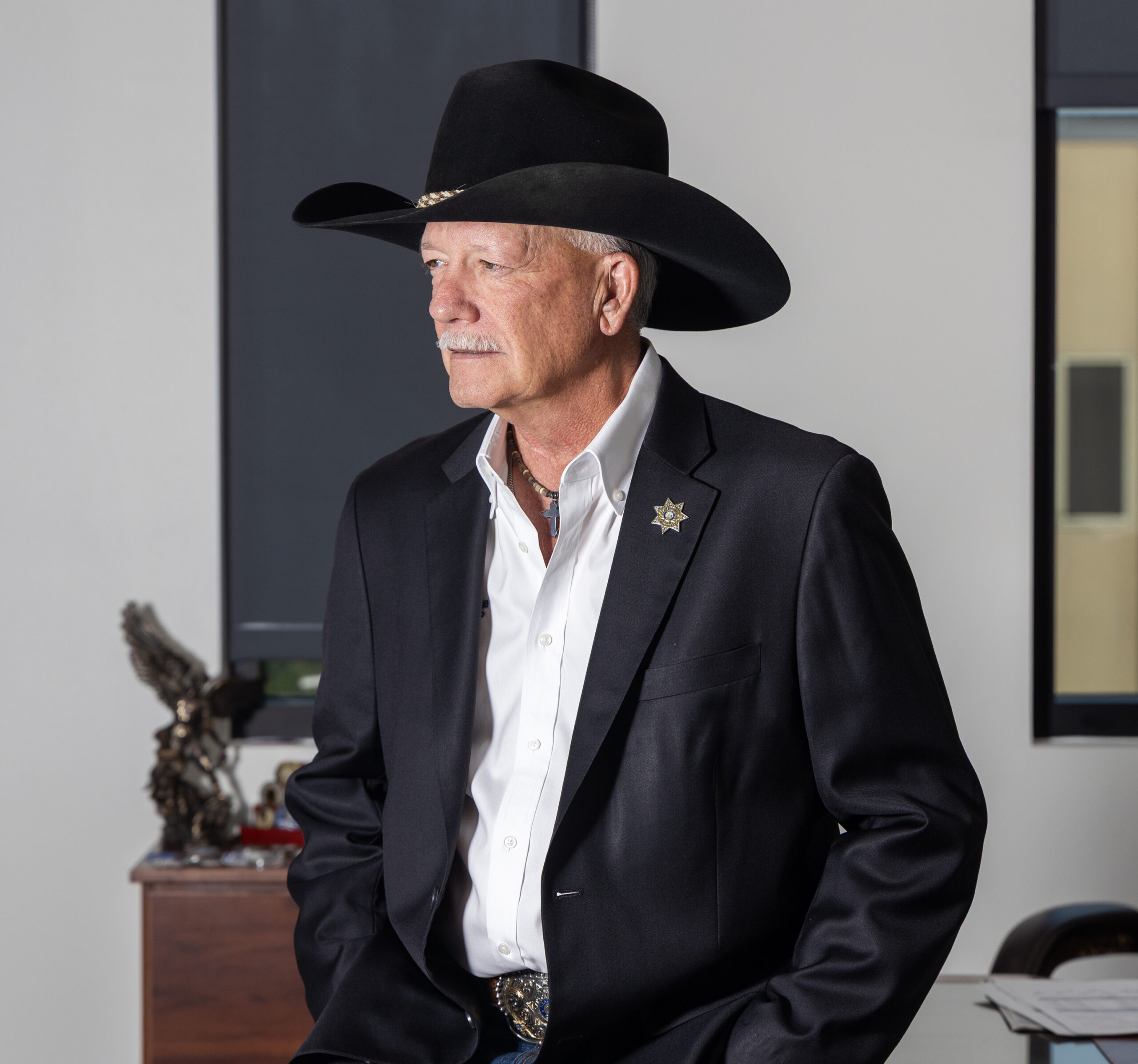 A man looks off frame toward his right in a room with vertical windows. He wears a black cowboy hat, a sheriff’s star pinned in the lapel of a black blazer, a large silver belt buckle and a silver cross on a necklace.