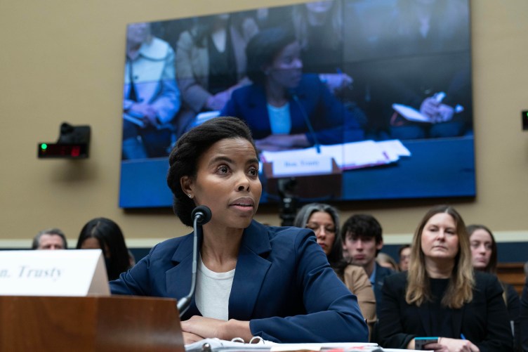 A woman in a blue suit sits at a table with a microphone in front of her. The same woman also appears on a large television screen on the wall behind where she is sitting.