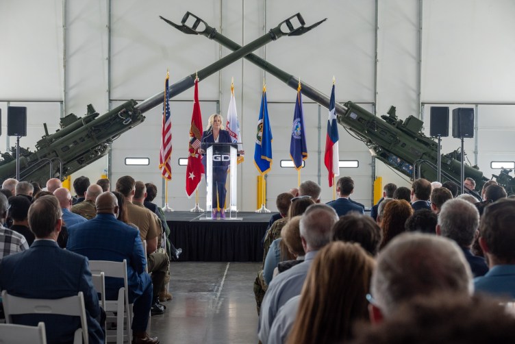 A woman addresses a crowd from a podium branded with the General Dynamics logo, on a stage displaying various flags and two howitzers.