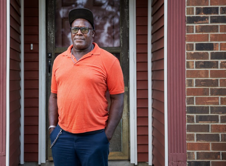A man wearing an orange polo shirt, silver chain necklace, baseball cap and yellow-tinted glasses looks into the camera. He is standing in front of a red-brick building with red siding.