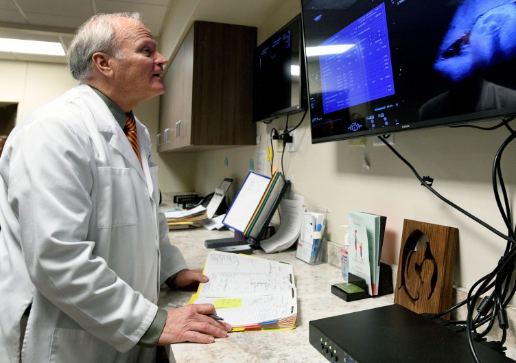 A man with white hair and wearing a white doctor’s coat stands and looks at a large monitor displaying medical scans.