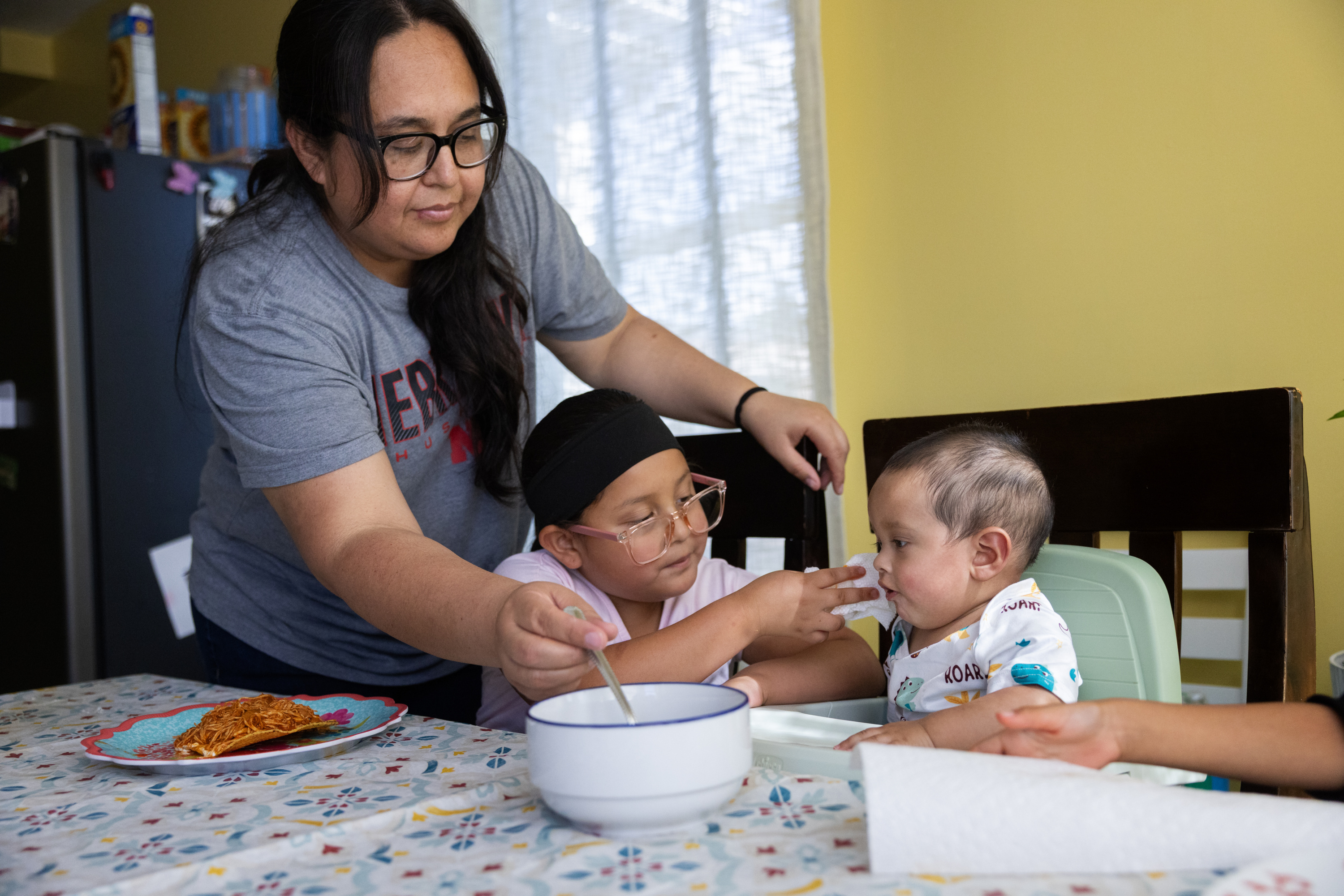 A woman with long black hair wearing a gray T-shirt and glasses stirs a spoon in a bowl of soup. She is leaning over a small child wearing pink glasses, a pink shirt and black headband. The child is touching a baby seated at the table. There is a yellow wall, window and refrigerator behind them. A plate of food is next to the woman’s arm on the table.