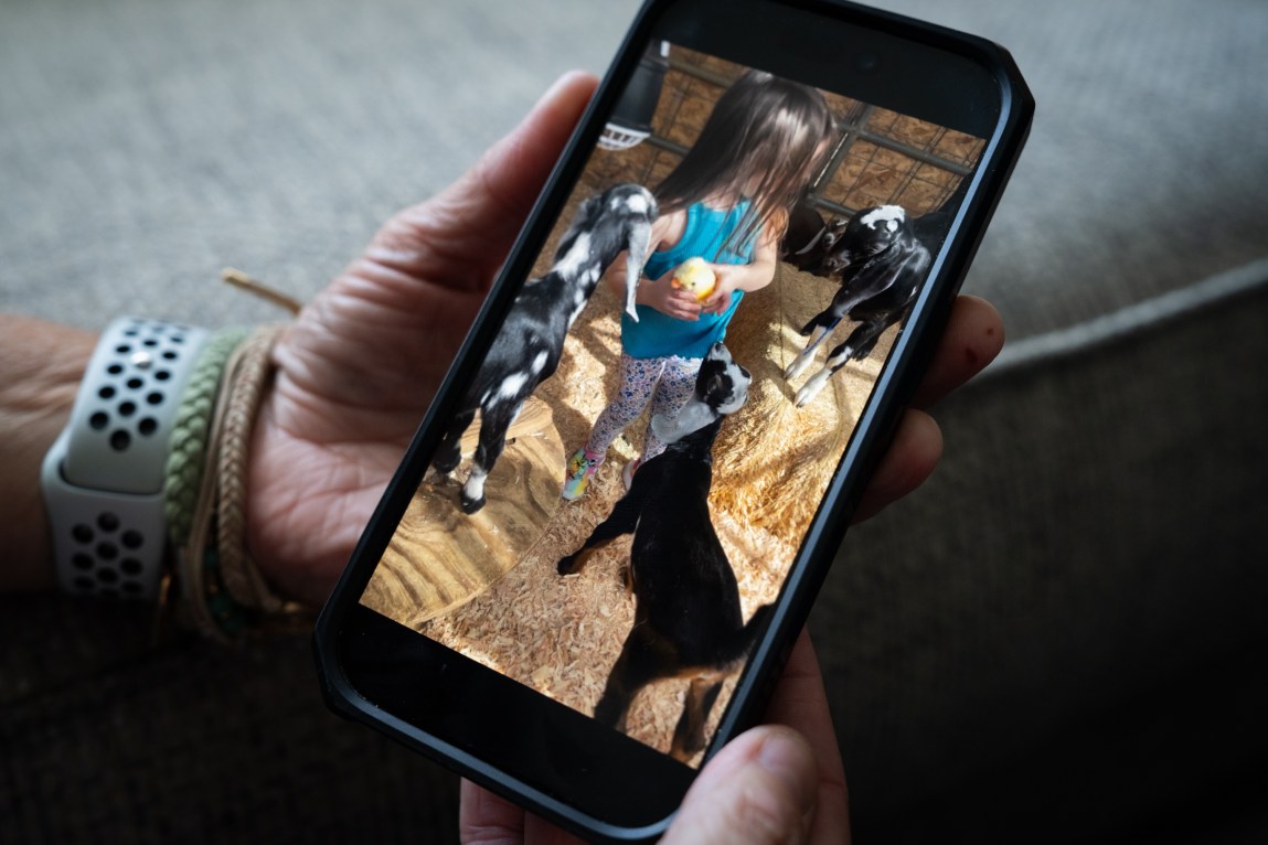 A close-up of a hand holding a smartphone displaying a photo of a young girl playing with baby goats in a hay-filled pen.