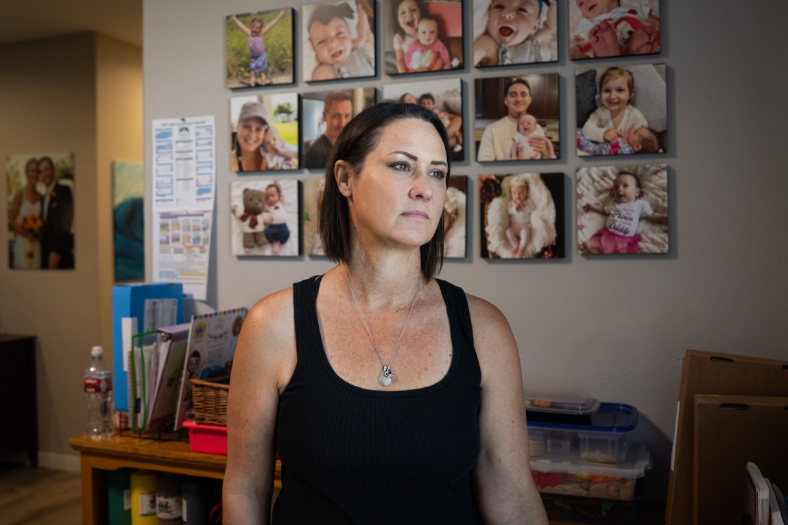 A woman with short, dark hair wearing a black tank top stands indoors with a serious expression, with a wall of framed family photographs behind her.