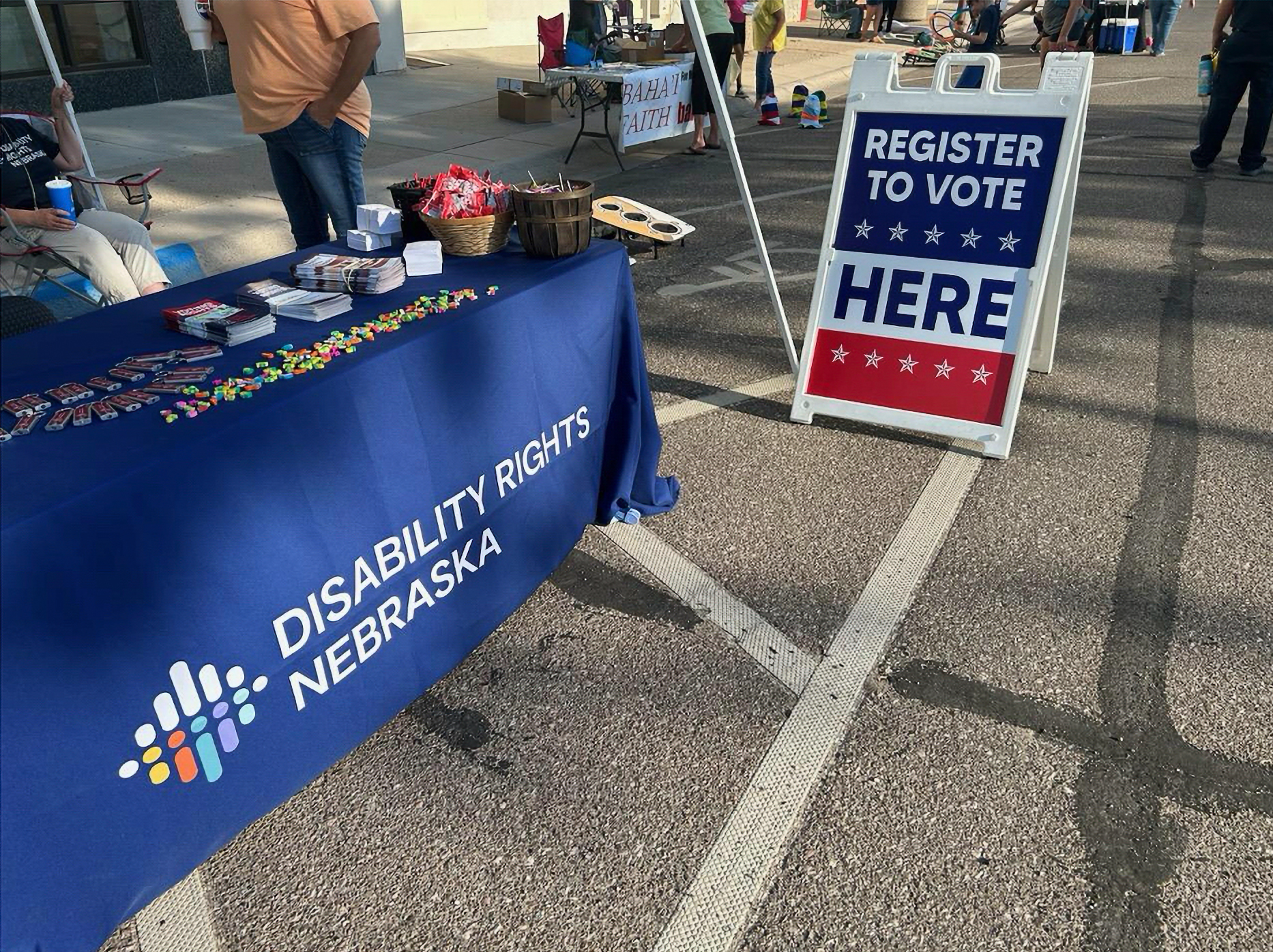 A table with a blue banner saying “Disability Rights Nebraska” sits on the left side of the image while a red, white and blue sandwich board directs people to register to vote at the table.