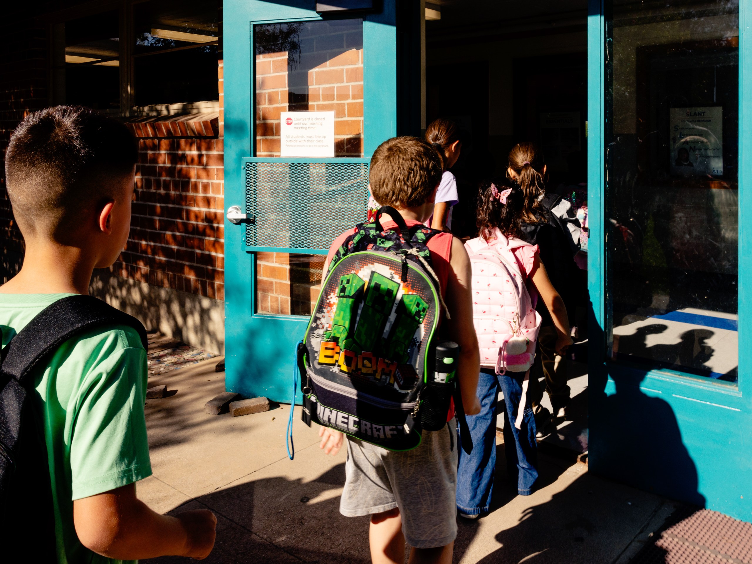 Several young students wearing backpacks head into a classroom on a bright, sunny morning.
