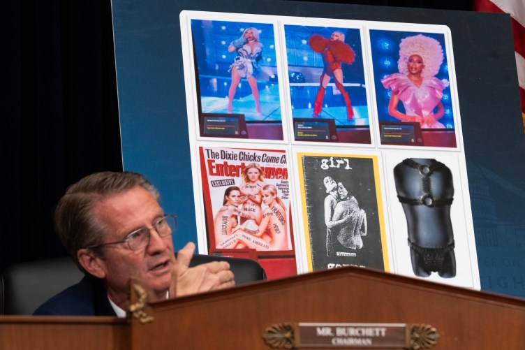 A man with glasses speaks at a podium in front of a poster showing drag queens, a leather harness and an Entertainment Weekly cover featuring the band the Dixie Chicks with derogatory remarks painted on their partially covered nude bodies.