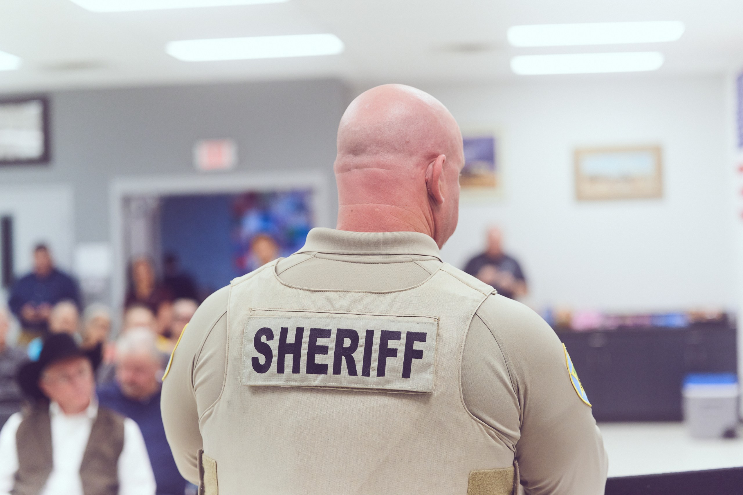 A bald man in a police uniform stands with his back toward the camera during a meeting in a bright room. A patch on his back says, “Sheriff.”