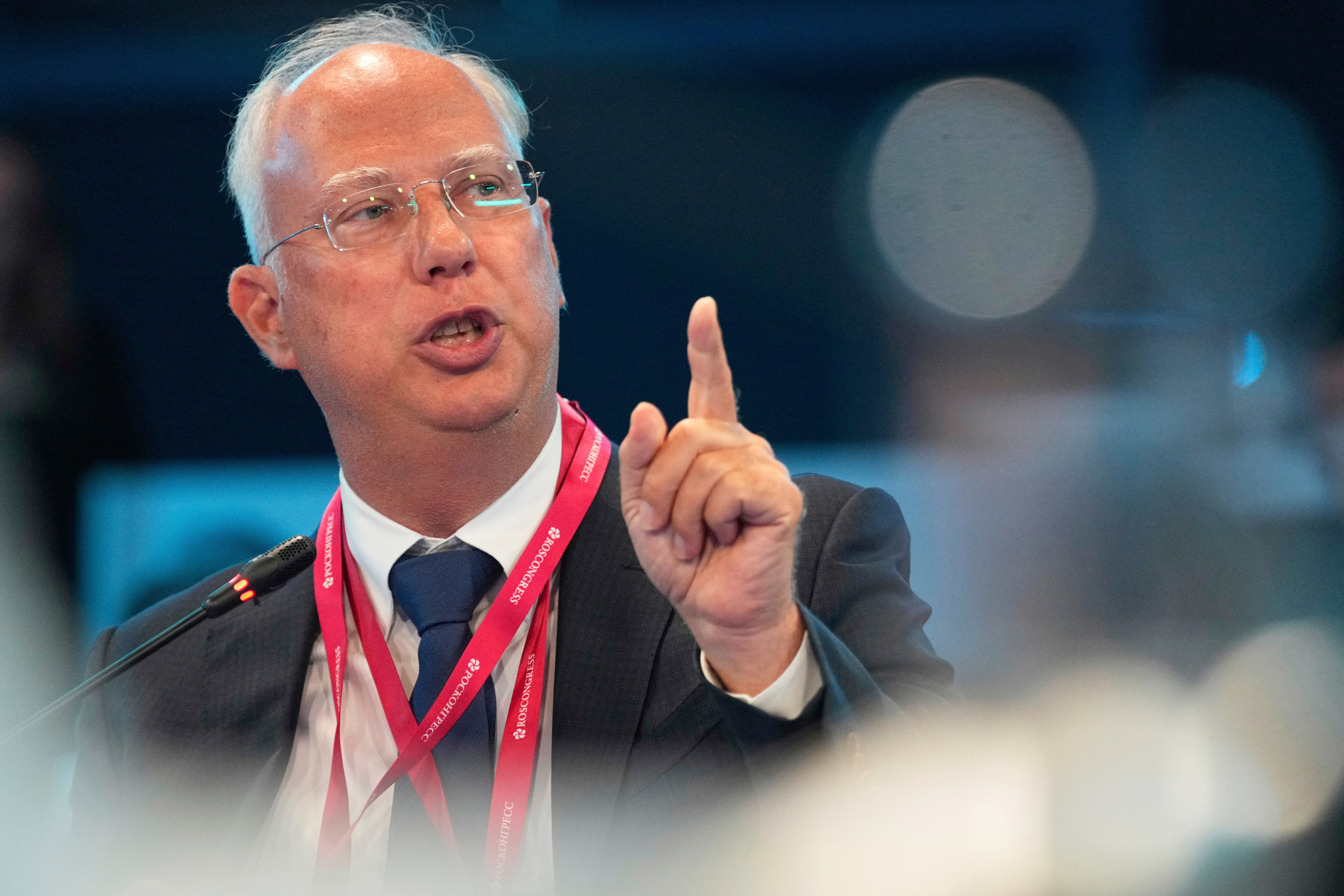 A man with white hair, glasses and two red lanyards around his neck holds up his left index finger and talks in front of a microphone.