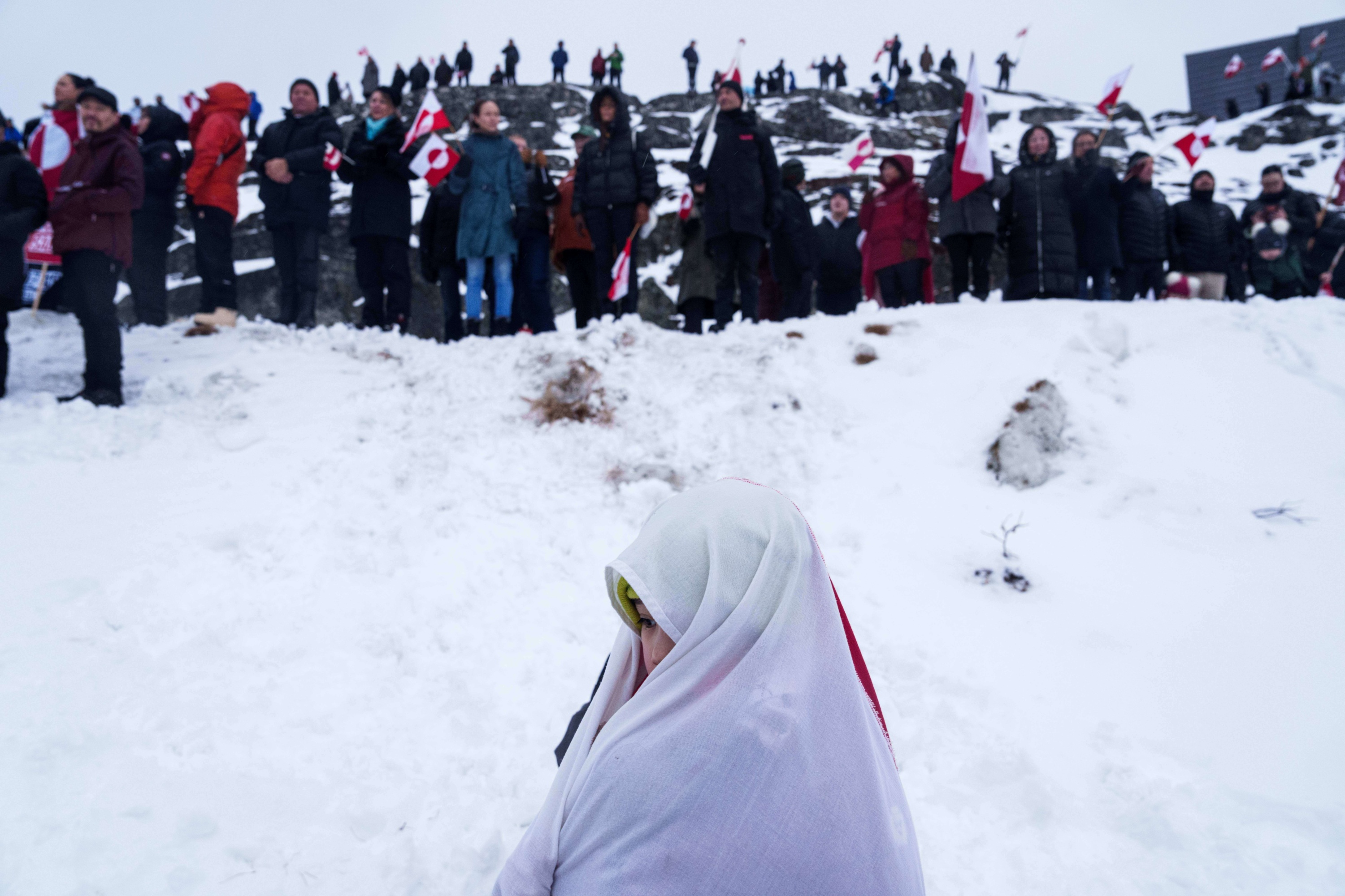 A person wrapped in a white cloth stands alone in front of a snowbank with many people standing holding Greenland’s flag.