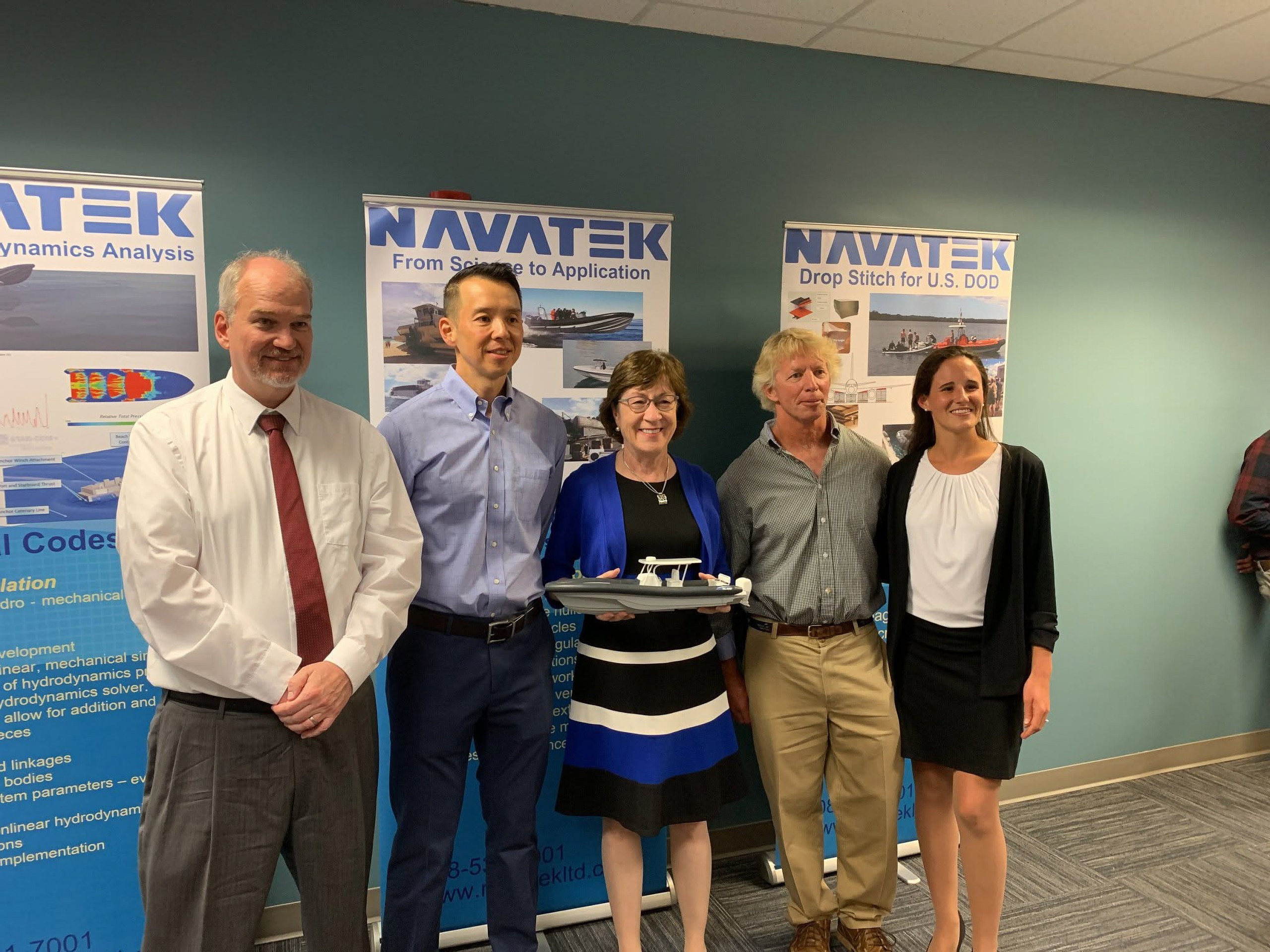 A group of three men and two women pose for a photo in front of banners for a defense contractor. The woman in the middle of the group holds a model of a boat in her hands.