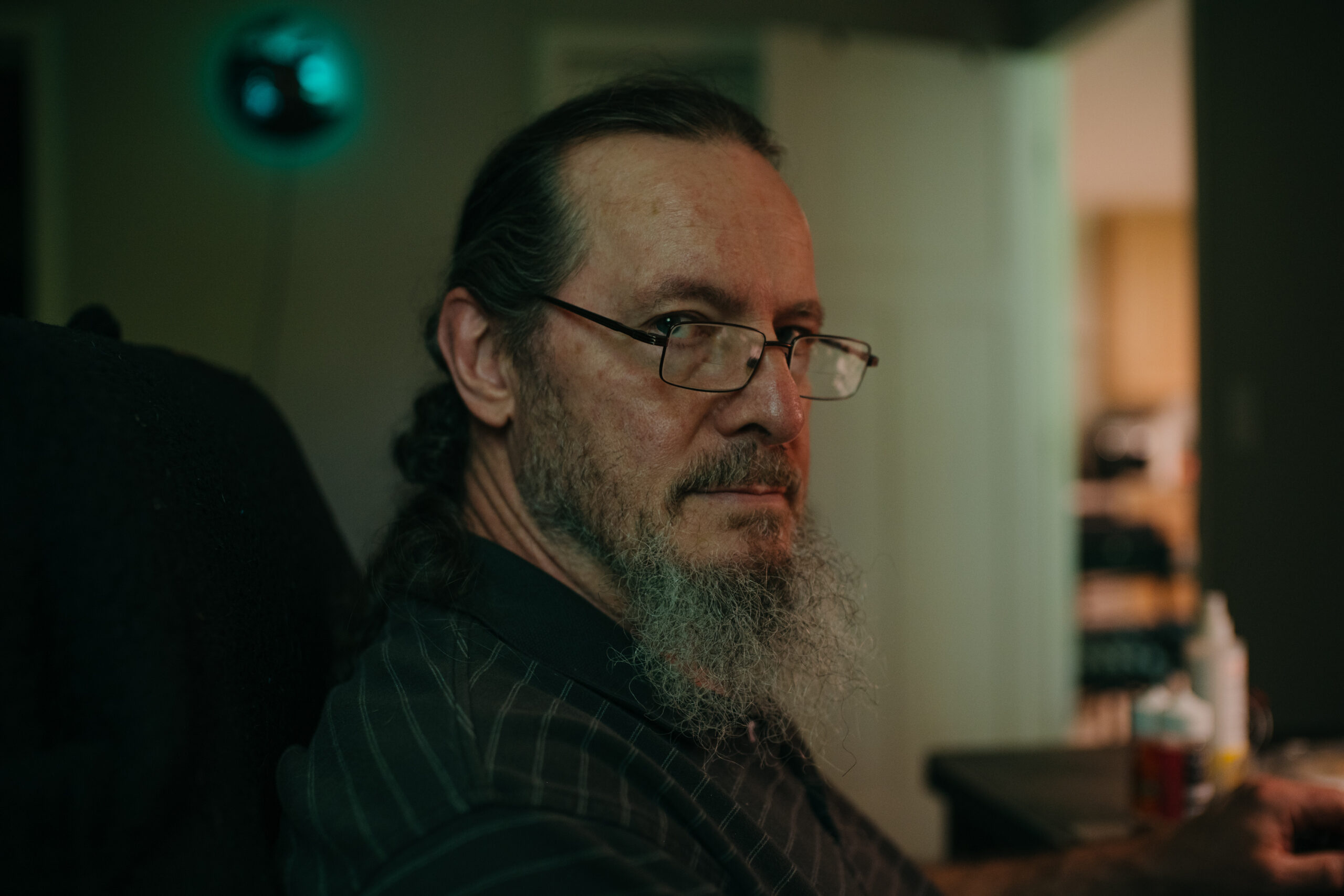 A man with a gray beard, ponytail and glasses sits inside a dimly lit room and stares at the camera.