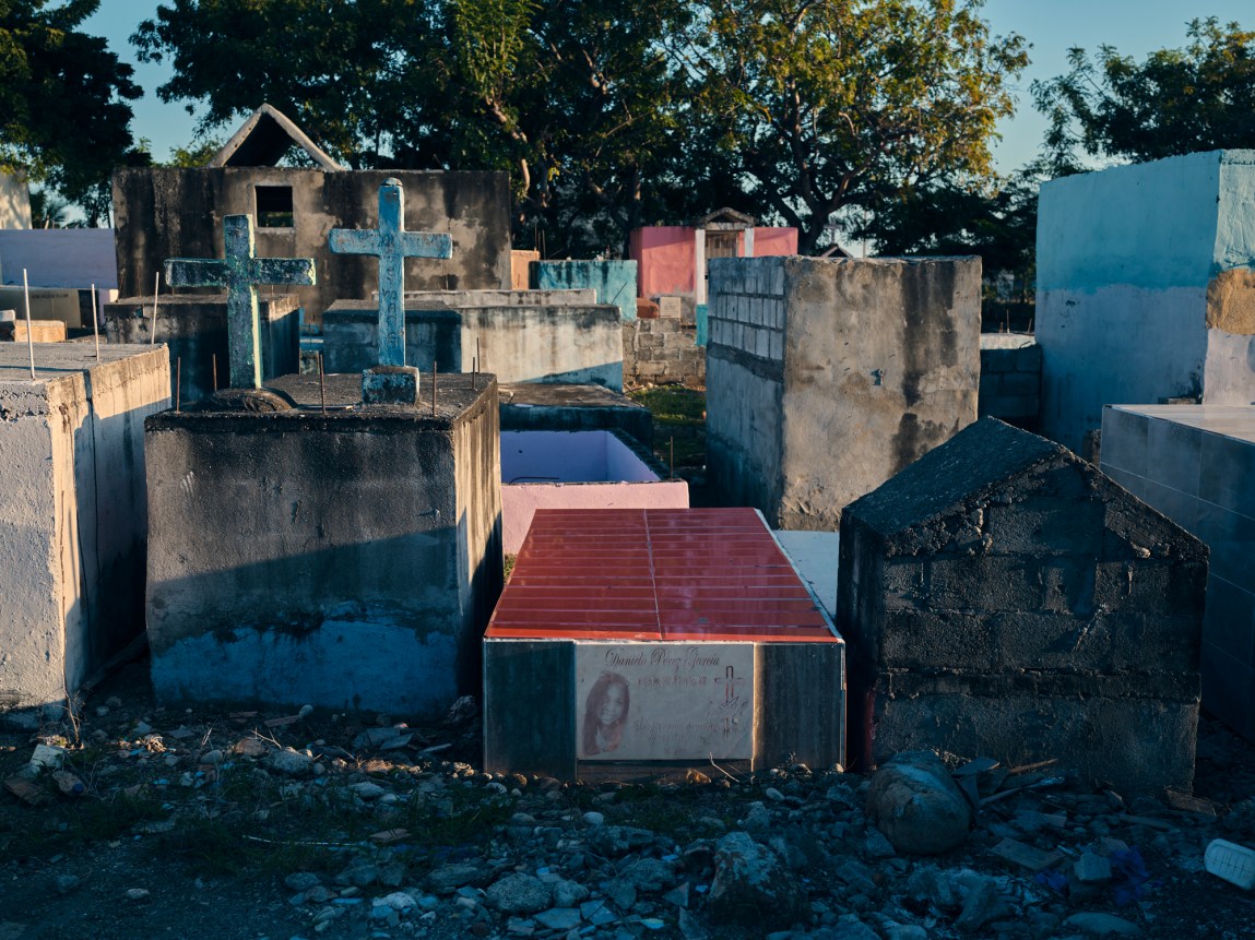 Concrete, box-shaped concrete tombs and stone crosses stand closely packed together in an outdoor cemetery under sunlight.