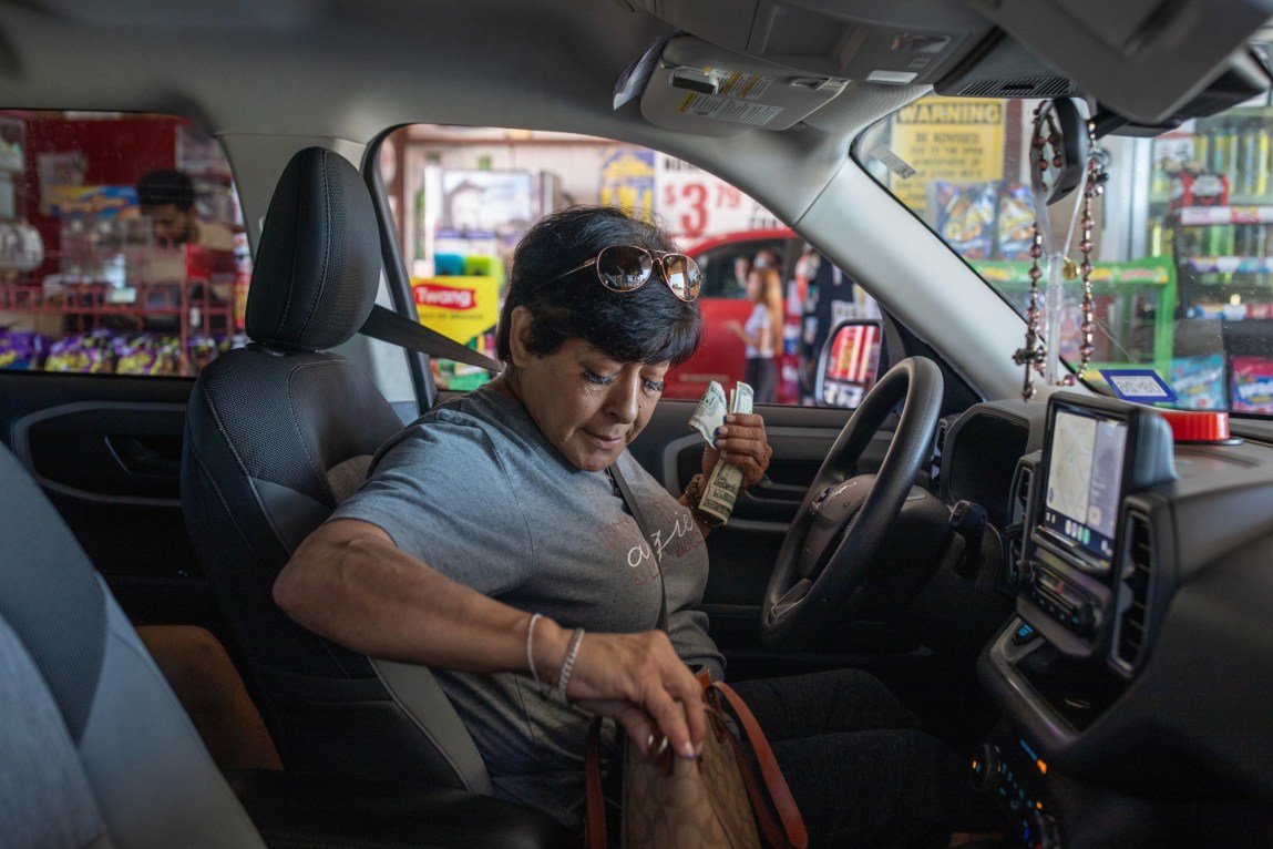 A woman sits in the driver’s seat of her vehicle, looking down at a purse as she holds money in her left hand. A rosary dangles from the rearview mirror.