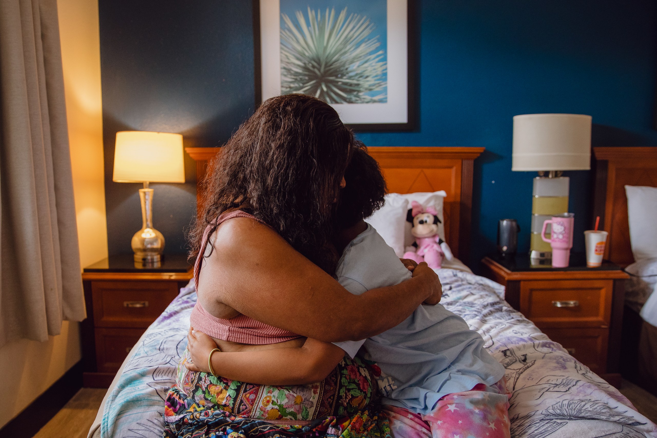 A woman and young child hug each other while sitting on the bed in a hotel room. Their hair obscures their faces, and the child is wearing pajamas.