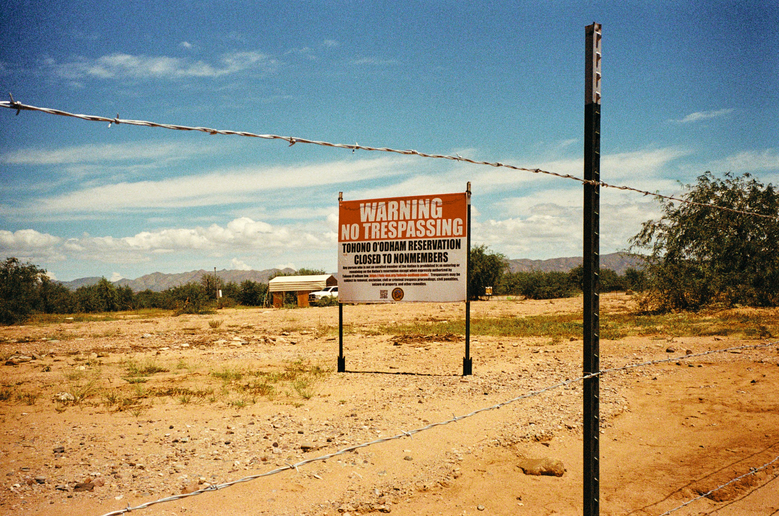 A red and white sign in a desert landscape is seen through a barbed-wire fence and warns against trespassing on the Tohono O’odham Nation for anyone who’s not a tribal member.