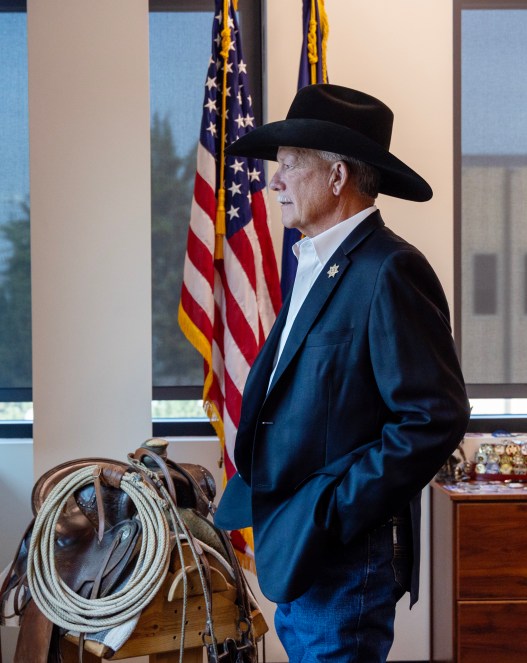 A man wearing a black cowboy hat, a navy blazer and jeans stands in profile to the camera, in front of a U.S. flag, a dresser of medals, a horses’ saddle and a row of windows.