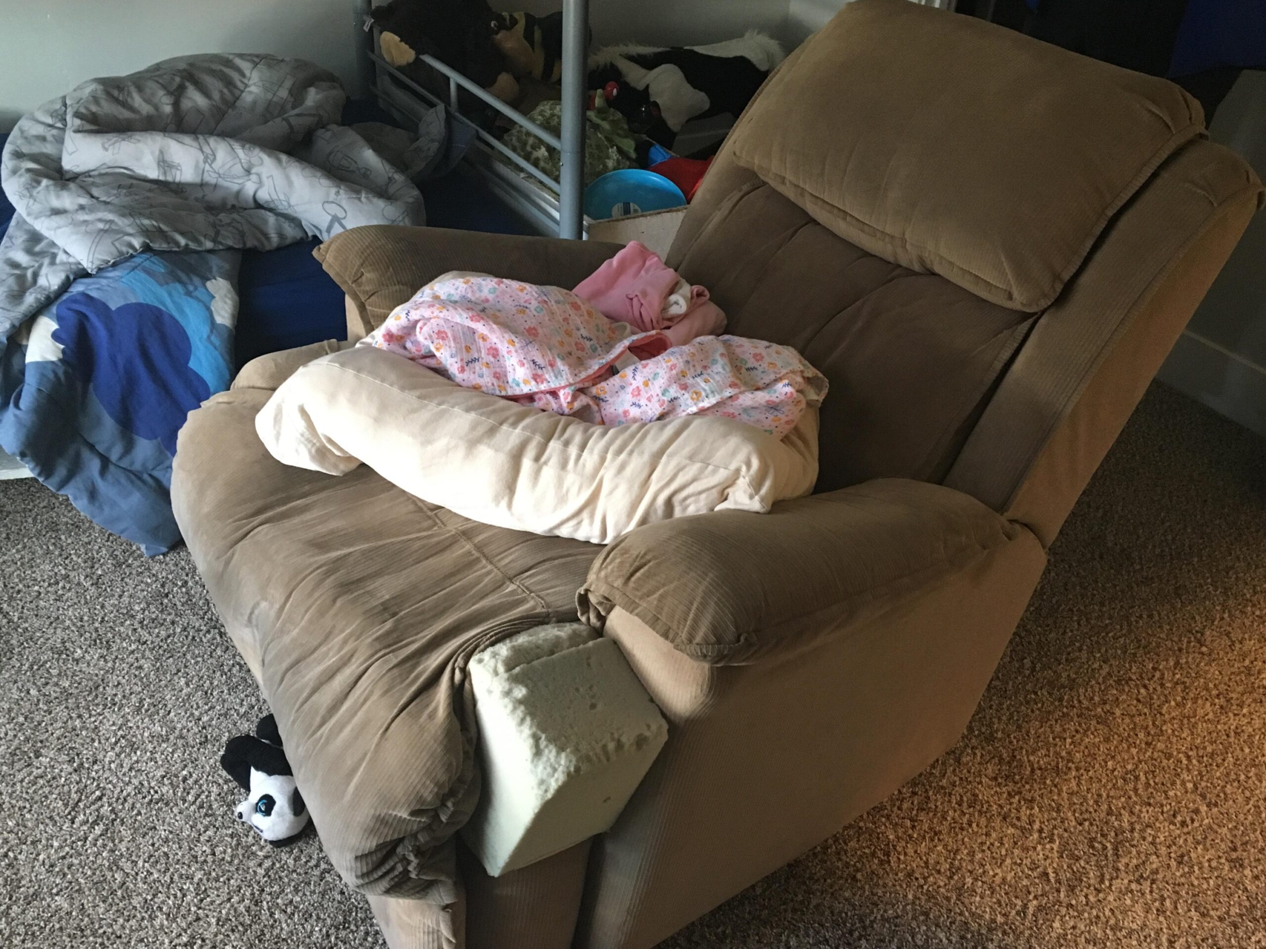 A bundle of pink blankets rests on a pillow on a brown recliner. A bedframe is behind the chair, next to a box of stuffed animals and toys. A small stuffed panda peeks out from below the chair.