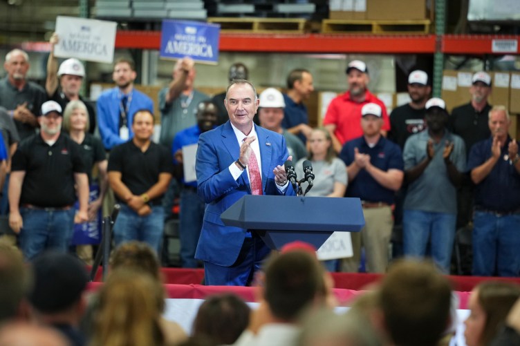 A man in a suit stands at a podium and claps his hands while a crowd of people behind him also clap.
