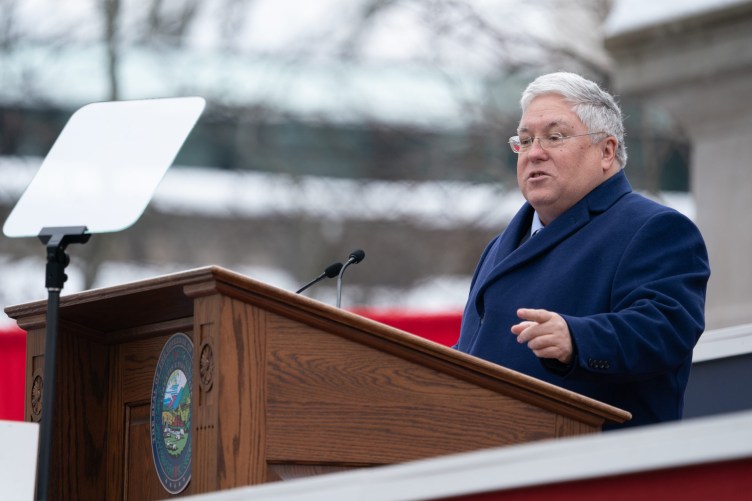 A man with white hair wearing a blue coat stands outside at a wooden lectern.