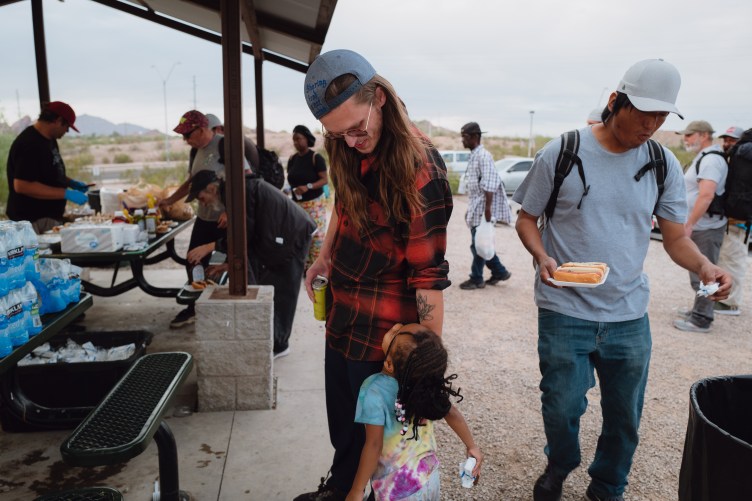 People gather around a picnic table at an outdoor park event offering free meals and water bottles to attendees.