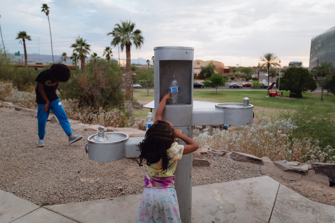 A young girl reaches up toward a water bottle set inside a public park hydration station while another child plays nearby.