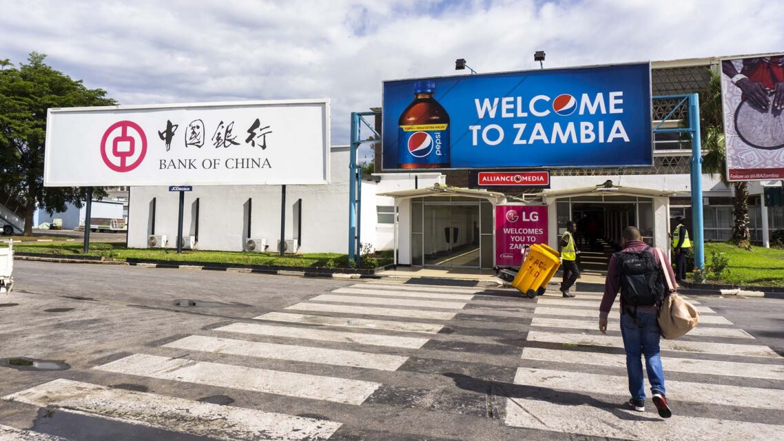 Photo of a man walking past a sign at the airport in Lusaka that reads Welcome to Zambia beside an advertisement for the Bank of China