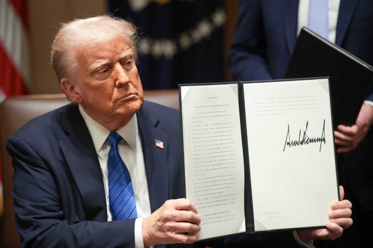 A man in a blue suit and tie sits at a table holding open a binder to display a signed executive order.