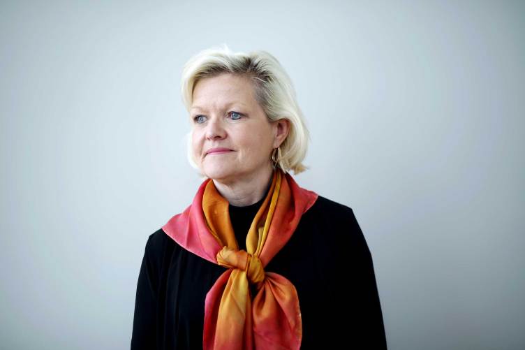 A portrait of a woman in a black shirt with a vibrant orange-and-yellow silk scarf tied around her neck, looking off to the side against a gray background.